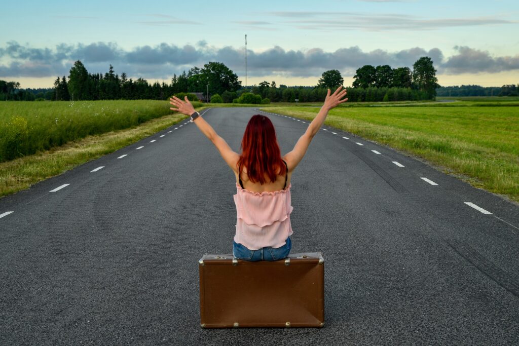 Red-haired woman sitting on a suitcase in the middle of a country road, arms raised in freedom.